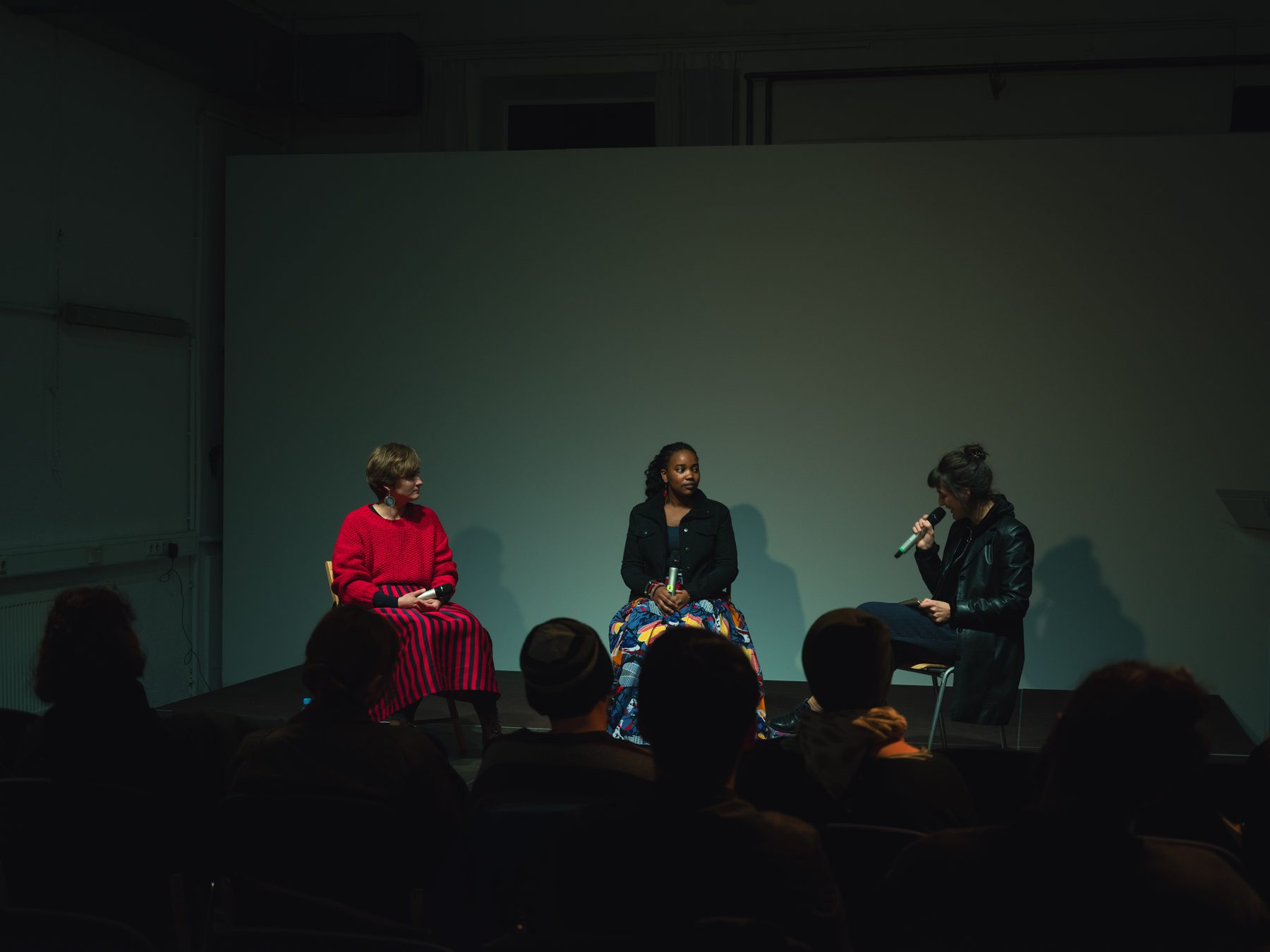 Three people seated on stage in conversation, one speaking into a microphone, in front of an audience