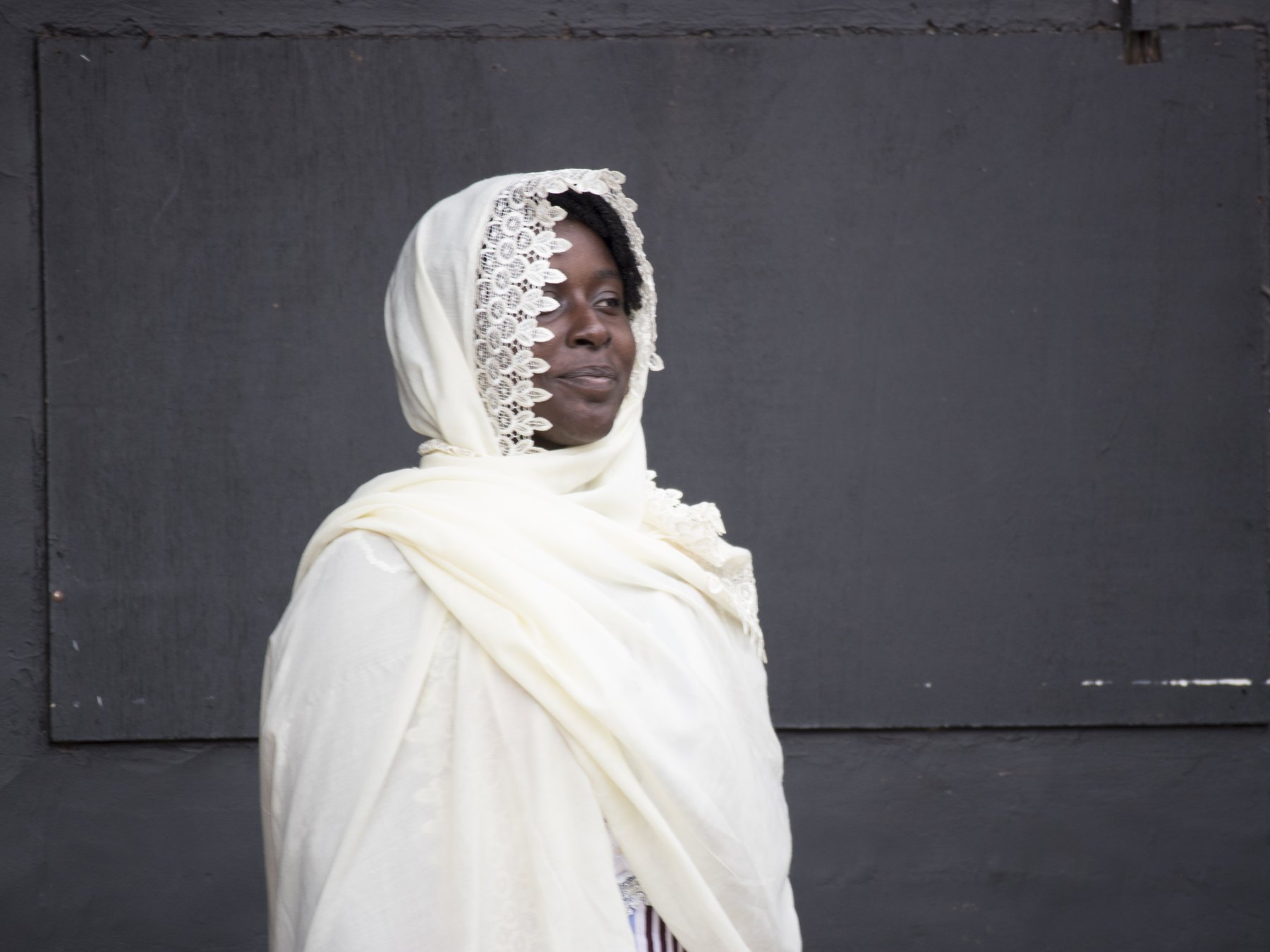 The Bride, in a white lace-edged veil and shawl, glances to one side against a dark wall