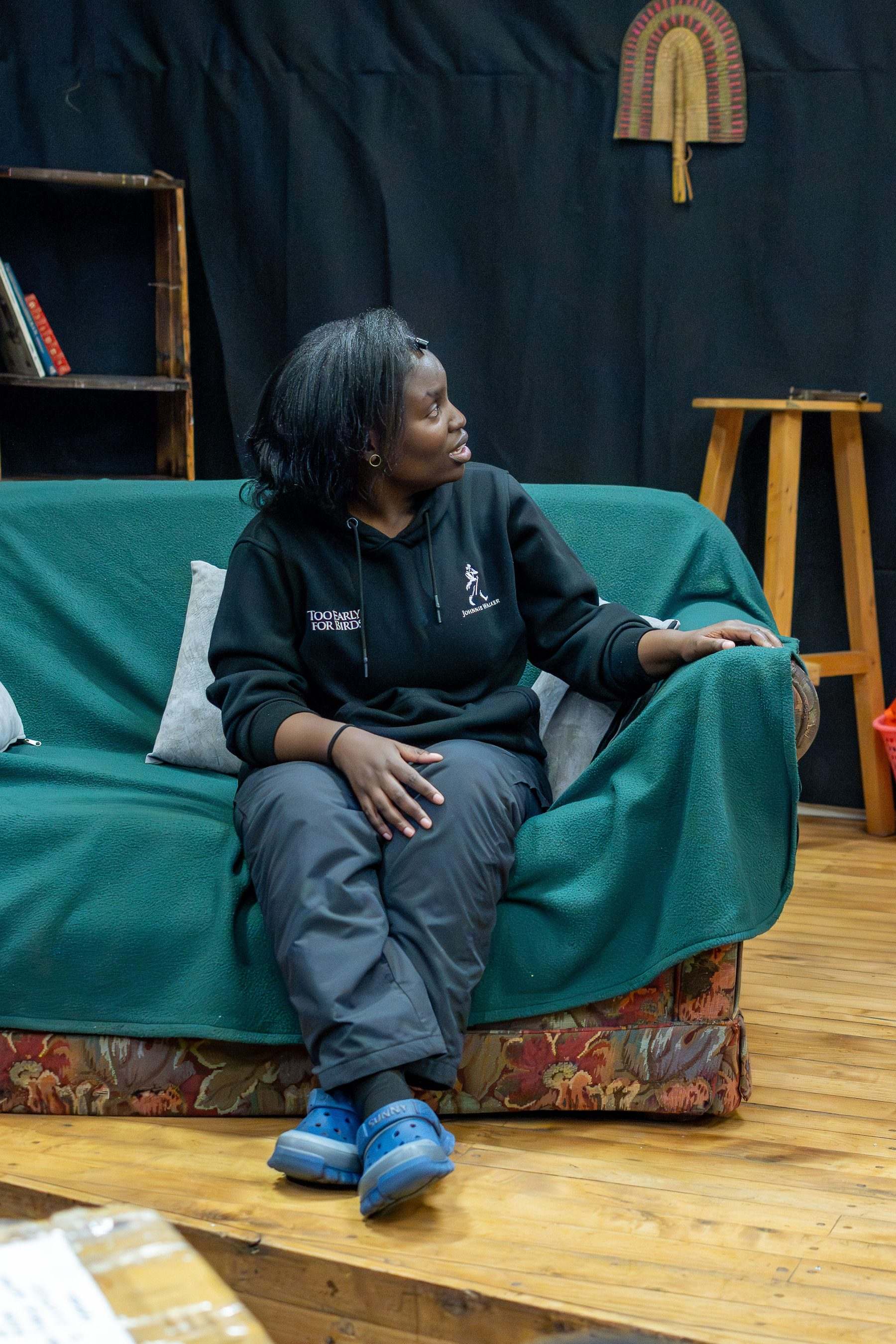 An actor sits on a green sofa on the set, turned to speak off to one side