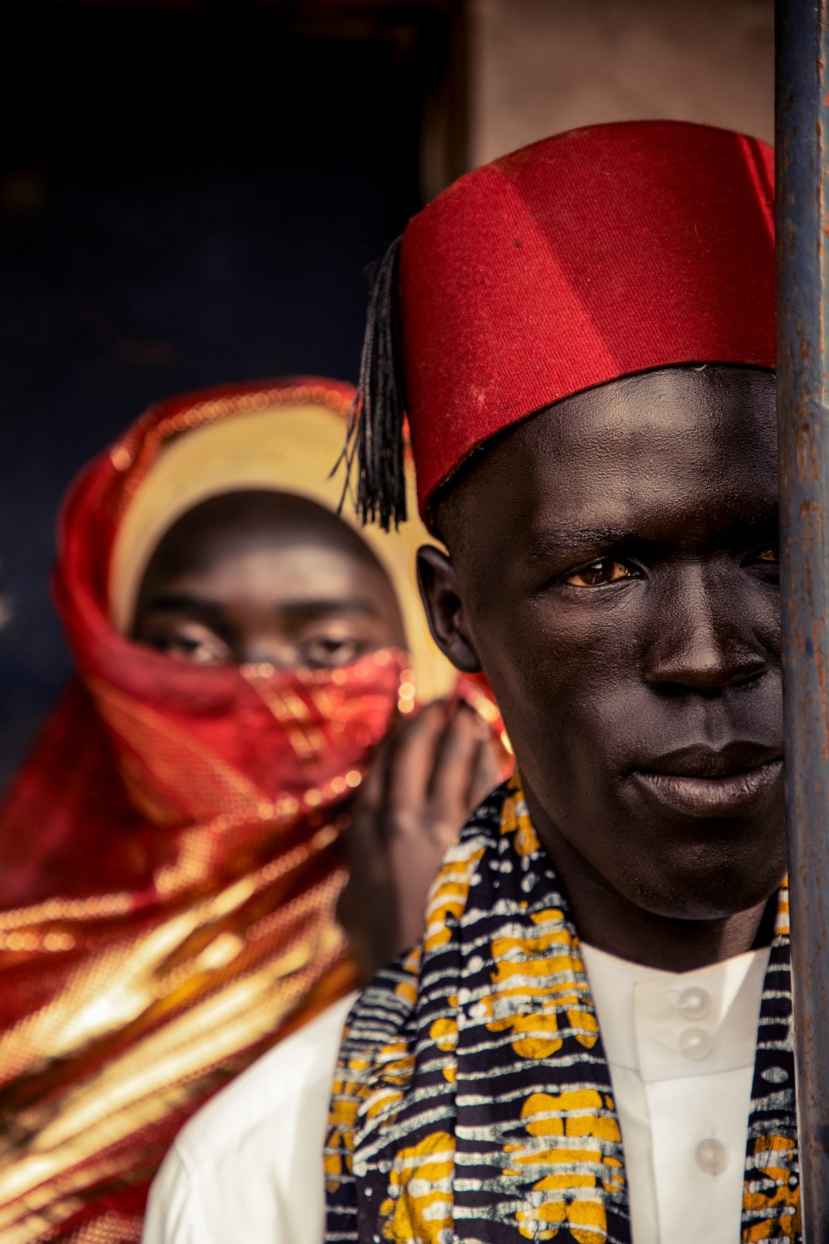 A man in a red fez looks into the camera; behind him, a woman covers her face with a red veil