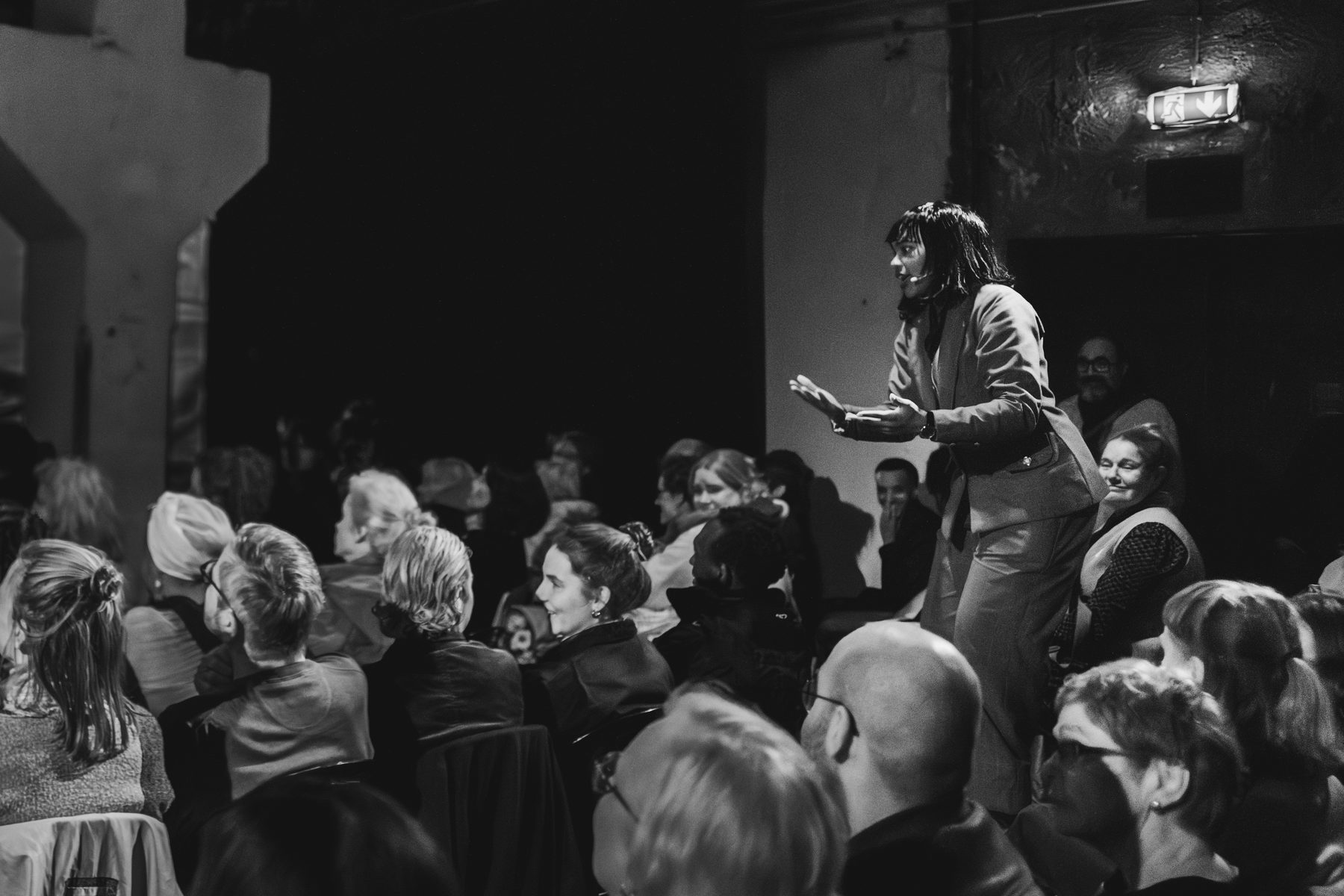 Black-and-white photograph of a performer standing among a packed audience, one hand raised as she speaks